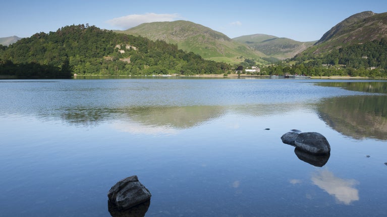 View across Ullswater at dawn, Aira Force and Ullswater, Cumbria, with hills in the background, their reflections in the water, and two rounded rocks near the shore